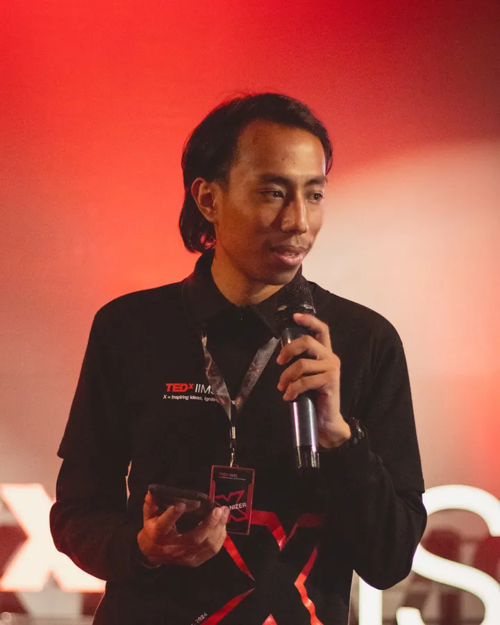 Aadarsh Shrestha speaking into a microphone on the TEDxIIMS stage, under red light.
